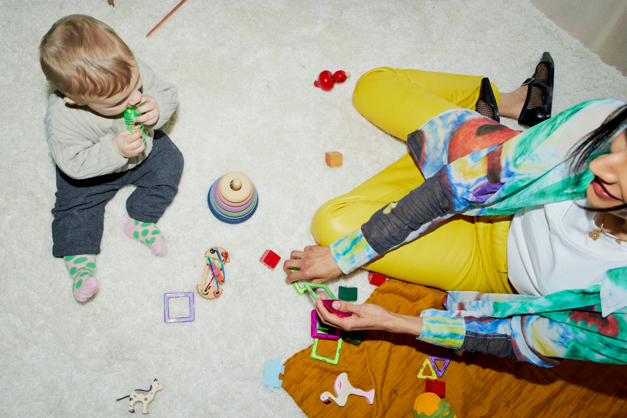 Baby on rug playing with colourful toys as adult helps — a safe, non-toxic playtime scene in a clean family home.
