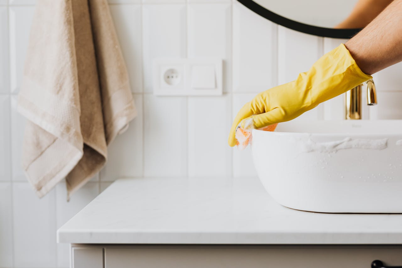 Hand in yellow glove scrubbing a bathroom sink with foam illustrating the debate between natural and chemical cleaners.