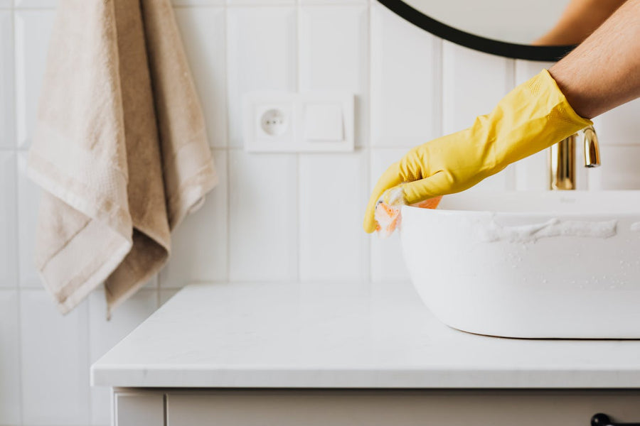 Hand in yellow glove scrubbing a bathroom sink with foam illustrating the debate between natural and chemical cleaners.